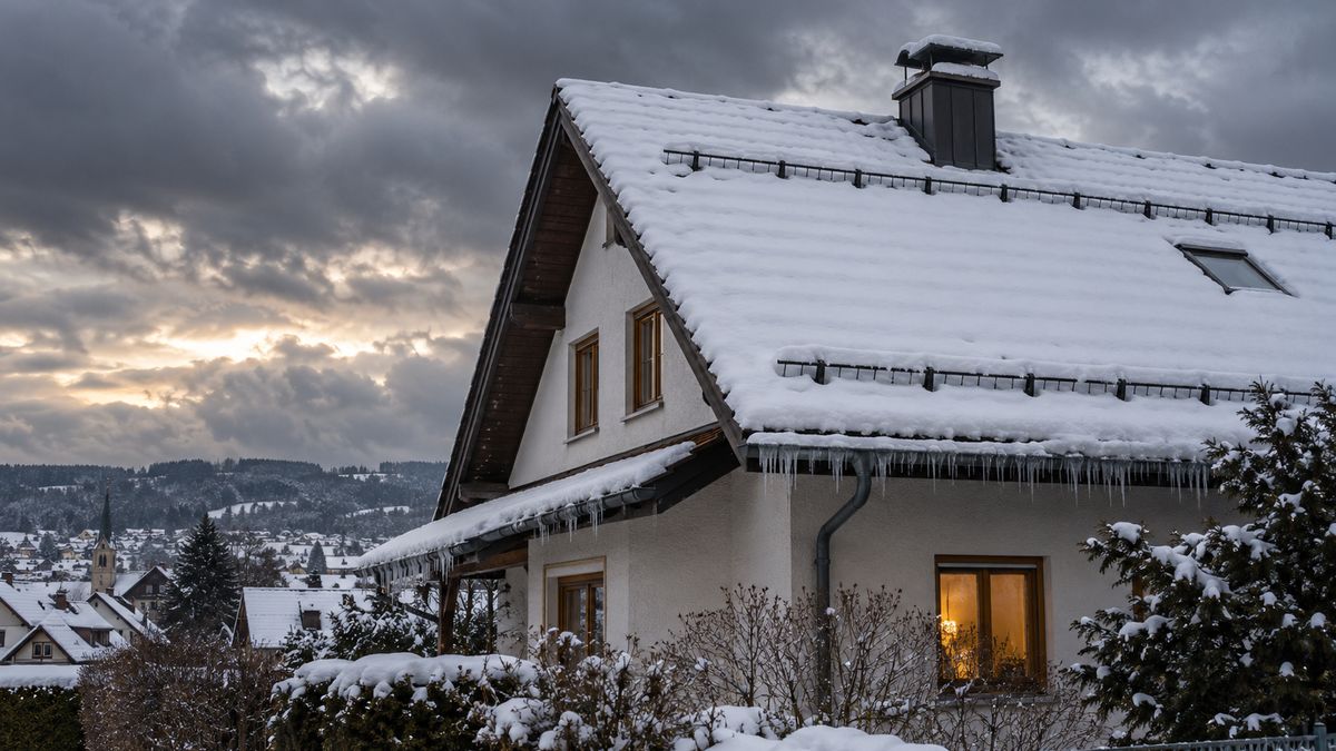 Schneebedecktes Satteldach mit Eiszapfen und Schneefanggittern vor stürmischem Winterhimmel